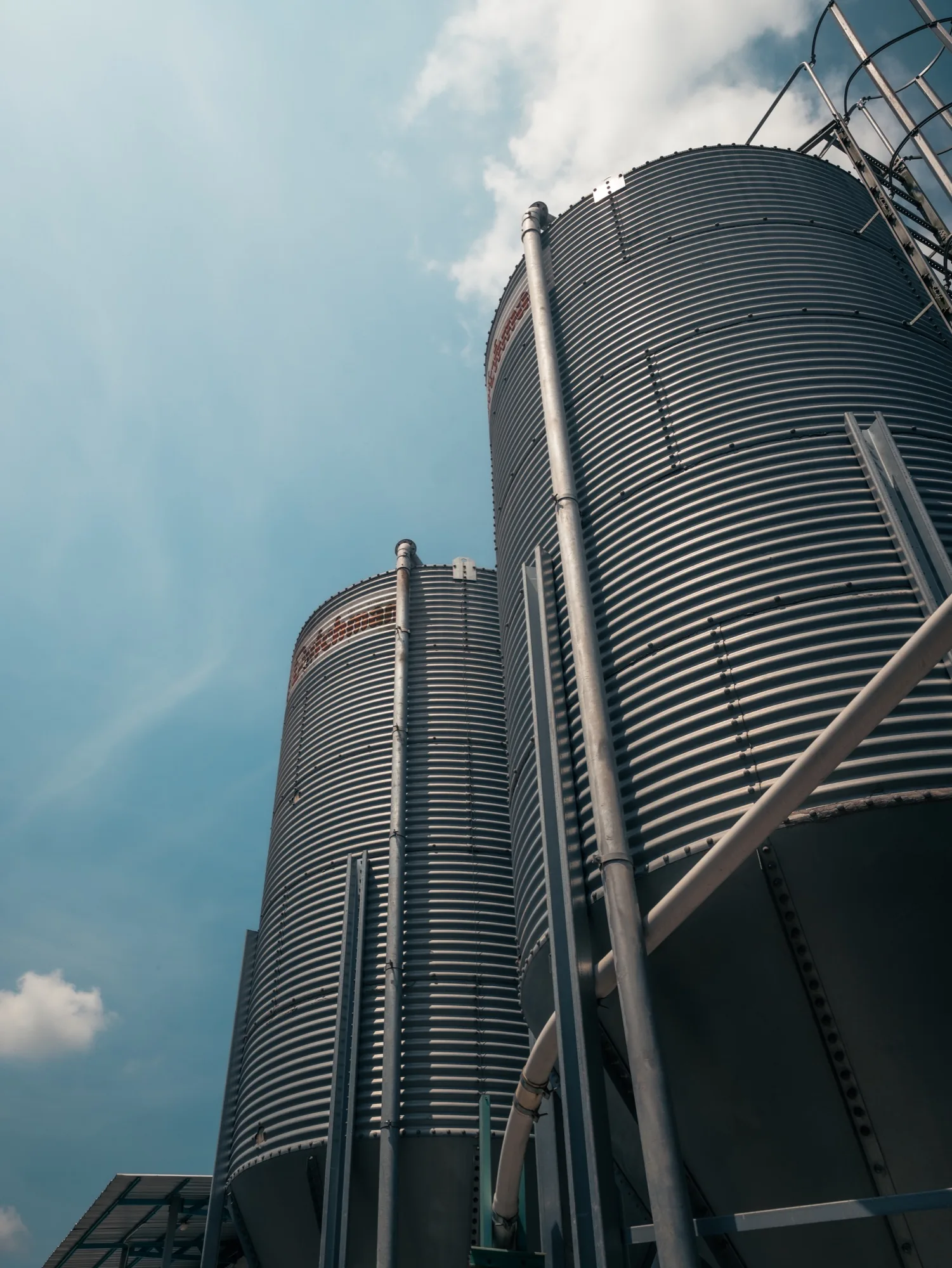 Low-angle view of corrugated metal feed silos at an agricultural farm facility, with metal pipes and support structures set against a blue sky.
